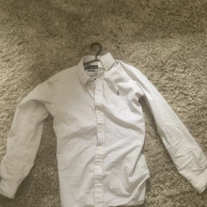 Vit och grå rutig skjorta från Ralph Lauren - Klassisk vit skjorta med grått rutmönster från Ralph Lauren. Skjortan har lång ärm, knappar framtill och broderad logga på bröstet. Tillverkad i bomull med stretch för extra komfort. Varan är nästan helt ny