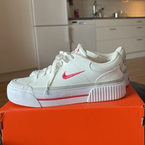 Vita Nike sneakers med rosa detaljer - Säljer ett par fräscha vita Nike sneakers med rosa detaljer. Skorna har en platt sula och är låga i modellen. Perfekta för dig som gillar sportig och stilren look.
