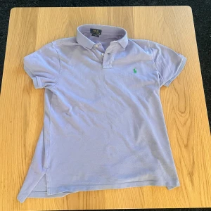 Ljuslila pikétröja från Polo Ralph Lauren - Säljer en ljuslila pikétröja från Polo Ralph Lauren med klassisk krage och knappar framtill. Tröjan har korta ärmar och en grön broderad logga på bröstet. Modellen är Custom Fit och passar perfekt till en avslappnad stil.