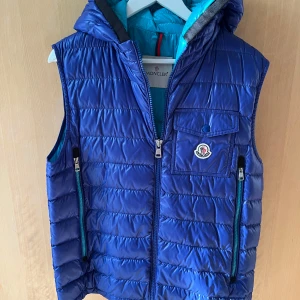 Blå dunväst från Moncler med huva - Snygg blå dunväst från Moncler med huva och dragkedja framtill. Västen har två fickor med dragkedja och en bröstficka med Moncler-logga. Insidan är turkos och västen är quiltad för extra värme.