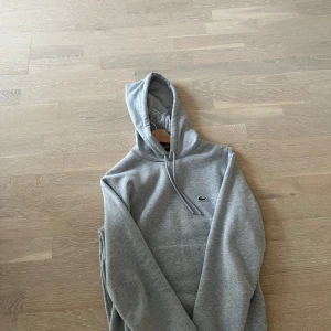 Grå hoodie från Lacoste - Säljer en klassisk grå hoodie från Lacoste med broderad logga på bröstet. Tröjan har huva med dragsko och långa ärmar. Perfekt för en avslappnad och stilren look.