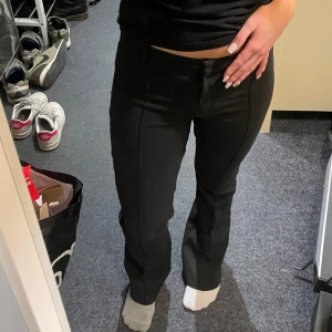 Svarta bootcut kostymbyxor - Säljer ett par svarta kostymbyxor med bootcut-snitt och låg midja. Byxorna har pressveck längs benen och klassiska hällor för bälte. Snygg passform som sitter tajt upptill och släpper ut vid benen. 