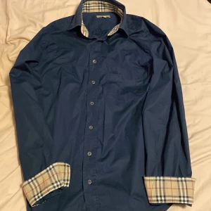 Mörkblå skjorta från Burberry med rutiga detaljer - Snygg mörkblå skjorta från Burberry i klassisk modell med rutiga detaljer på insidan av krage och ärmslut. Skjortan har knappar framtill och är tillverkad i bomull med lite stretch för extra komfort. 
