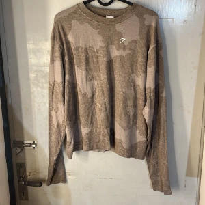 Tröja  från gymshark  - Säljer en beige långärmad tröja från Gymshark med camo-mönster och rund hals. Perfekt för träning eller chill dagar. Mjukt och lätt material som andas. Gymshark-logga på bröstet.