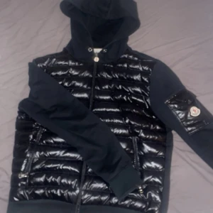 Moncler cardigan - Säljer en svart pufferjacka från Moncler med glansig framsida och ärmficka med logga Jackan har huva☀️, dragkedja och quiltade partier framtill. Ärmarna är i mjukare material och jackan har fickor med dragkedja.🏖️
