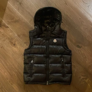 Svart dunväst från Moncler - Snygg svart dunväst från Moncler med huva och dragkedja. Västen har två fickor med dragkedjor och ett broderat Moncler-märke på bröstet. Perfekt för kyligare dagar.