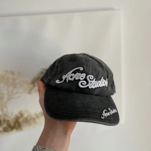 Svart keps från Acne Studios - Snygg grå keps från Acne Studios med vit broderad logga framtill och på skärmen. Justerbar rem baktill för perfekt passform. Klassisk och stilren design som passar till många outfits.