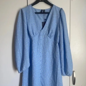 Ljusblå blommig klänning med v-ringning från Vero Moda - Supersöt ljusblå klänning från Vero Moda med små vita blommor, v-ringning och långa puffärmar. Klänningen har en markerad midja och lätt struktur i tyget. Perfekt för vår och sommar!