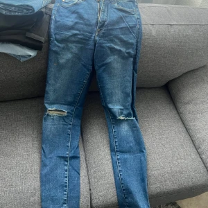 Blå skinny jeans med slitningar - Säljer ett par blå skinny jeans med slitningar på knäna och klassisk femficksdesign. Jeansen har hög midja och dragkedja med knapp framtill. Perfekta för en avslappnad och trendig look. JEANSEN har två fläckar som kanske går bort i tvätten 