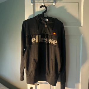 Svart hoodie från Ellesse - Säljer en svart hoodie från Ellesse med dragkedja och huva. Stor logga framtill i vitt och klassiska Ellesse-detaljer. Tröjan har långa ärmar och två fickor framtill. Perfekt för en sportig och avslappnad stil.
