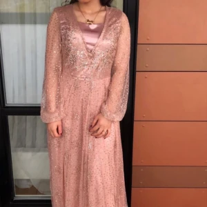 Glittrig rosa långklänning med v-ringning - Säljer en superfin rosa långklänning med glittriga detaljer och v-ringning. Klänningen har långa, transparenta ärmar och ett elegant fall. Perfekt för bal eller festliga tillfällen. Materialet känns lätt och skimrande.