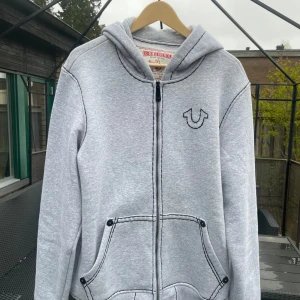 Grå hoodie från True Religion - Säljer en grå hoodie från True Religion med dragkedja och stor logga på ryggen. Tröjan har huva, två fickor fram och svart konstrastsömmar. Storlek M med loose passform. Köpt direkt i USA. 😁😁🙌