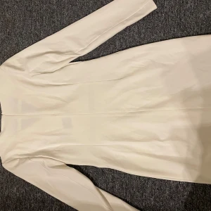 Vit långärmad klänning från Boohoo - Säljer en stilren vit klänning från Boohoo med lång ärm och v-ringad omlottkrage. Klänningen har fickor framtill och en figurnära passform som ger en snygg siluett. Perfekt för dig som vill ha en clean och modern look.