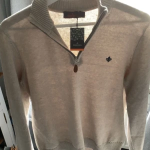 Beige halvzip-tröja från Morris - Säljer en stilren beige stickad tröja från Morris med halv dragkedja och hög krage. Tröjan har ett diskret broderat emblem på bröstet och ribbade muddar vid ärmslut och nederkant. Perfekt för lager-på-lager och snygg till jeans.