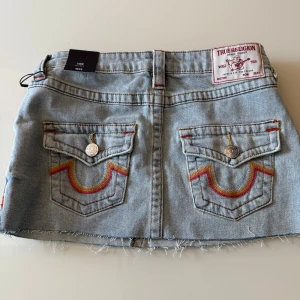 Ljusblå jeanskjol från True Religion - Säljer en ljusblå jeanskjol från True Religion med rå kant nertill och klassiska bakfickor med färgglada broderier. Kjolen har knapp- och dragkedjestängning samt detaljerade ficklock med metallknappar.