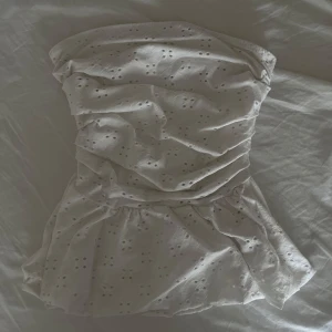 Vit axelbandslös tubtopp med broderade detaljer och volanger  - Säljer en söt vit tubtopp med broderade blommönster och volang nertill. Toppen är axelbandslös, perfekt för varma dagar. (Elastiskt band vid brösten)pris går att diskutera!