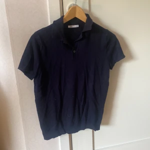Mörkblå pikétröja från Zara - Säljer en klassisk mörkblå pikétröja från Zara. Tröjan har korta ärmar, krage och knäppning framtill. Perfekt för en stilren och enkel look. Tillverkad i mjukt material som känns skönt mot huden.