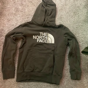 Mörkgrön hoodie från The North Face - Säljer en mörkgrön hoodie från The North Face med stor vit logga på bröstet. Tröjan har huva, magficka och ribbade muddar vid ärmslut och nederkant. Perfekt för en avslappnad stil.