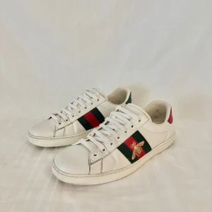 Vita sneakers från Gucci ace bee - Säljer ett par vita sneakers från Gucci med gröna och röda ränder på sidorna samt en broderad guldfärgad bi. Skorna har snörning och röd/grön detalj på hälen. Klassisk och stilren modell!
