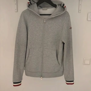 Grå hoodie från Moncler - Säljer en stilren grå hoodie från Moncler med dragkedja framtill och huva. Tröjan har långa ärmar och fickor på sidorna samt Moncler-logga på ärmen. Muddarna har diskreta ränder i rött, vitt och blått.