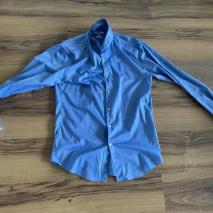Blå slim fit skjorta - Säljer en klassisk blå skjorta i slim fit-modell. Skjortan har långa ärmar och knäppning framtill med vita knappar. Perfekt för dig som gillar en stilren och enkel look.