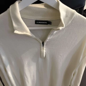 Vit stickad tröja med dragkedja från J.Lindeberg - Säljer en stilren vit stickad tröja från J.Lindeberg med halv dragkedja framtill och klassisk krage. Perfekt för en snygg och avslappnad look. Tröjan har långa ärmar och diskret logga på bröstet.