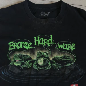 Bronze 56K - Svart t-shirt från Bronze 56K med tryck av tre grodor och texten 'Bronze... Hard... Ware' i grönt. Klassisk passform och rund halsringning. Perfekt för dig som gillar unika och roliga motiv. Storlek XL.