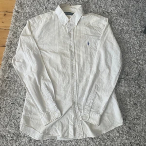 Vit skjorta från Ralph Lauren - Klassisk vit skjorta från Ralph Lauren med broderad logga på bröstet. Skjortan har lång ärm, knappar framtill och krage. Perfekt för en stilren och tidlös look.