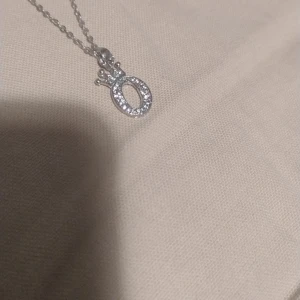 Silverfärgat halsband med krona och stenar - Säljer ett elegant halsband i silverfärg med ett O-format hänge dekorerat med små glittrande stenar och en liten krona upptill. Perfekt för dig som gillar stilrena smycken med en touch av glamour. Pris kan diskuteras.
