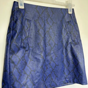 Blå ormmönstrad kjol från NA-KD - Säljer en blå kortkjol från NA-KD med ormmönster och två fickor fram. Kjolen har dragkedja bak och är tillverkad i ett syntetmaterial med lätt glansig finish. Perfekt statement-plagg för dig som gillar djurmönster.