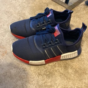 Adidas NMD R1 sneakers i marinblått - Säljer ett par Adidas NMD R1 sneakers i marinblått med röda och vita detaljer på sulan. Skorna har klassiska tre ränder på sidan, snörning och mesh-ovandel för extra komfort. Perfekta för dig som gillar streetwear och sportig stil. Defekt men inget som man tänker på