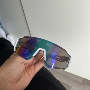 Vita sportiga solglasögon med spegelglas - Säljer ett par sportiga solglasögon med vit ram och stora spegelglas i blått och grönt. Glasögonen har en futuristisk design med bred båge och passar perfekt för träning eller utomhusaktiviteter.