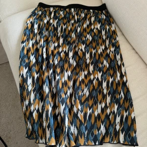 Mönstrad kjol från Vila - Snygg plisserad kjol från Vila med grafiskt mönster i blått, vitt, svart och senapsgult. Kjolen har resår i midjan och är i ett luftigt och lätt material. 