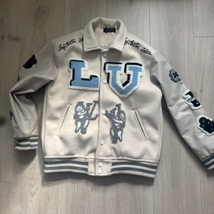 Beige och grå varsityjacka med broderade detaljer - Säljer en beige och grå varsityjacka från  med stora broderade bokstäver och figurer i blått, svart och vitt. Jackan har ribbade muddar, knappar framtill och flera patchar på ärmar och rygg. Perfekt statement-plagg för dig som gillar streetwear. Längd 64 cm. Storlek M passar S. Har ett litet hål inne. Men går att sy 