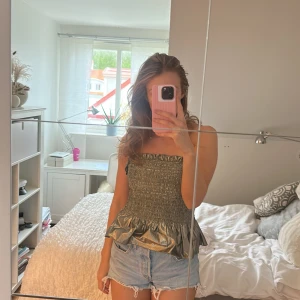  glansig tubtopp med volang - Säljer en snygg grön tubtopp med glansigt material och volang nertill. Toppen är smockad över bysten och har en croppad passform. Perfekt för dig som vill sticka ut lite extra!