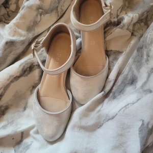 Glittriga pumps med ankelrem - Säljer ett par ljusa pumps från Poppe med glittrig finish och justerbar ankelrem med guldfärgat spänne. Skorna har låg klack och rund tå, perfekta för dig som vill ha något stilrent men ändå lite extra.