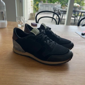 Valentino Rockrunner sneakers i svart och grått - Säljer ett par snygga Valentino Rockrunner sneakers i svart mesh och mocka med grå och vinröda detaljer. Skorna har snörning, rund tå och en unik nitudsdetalj på hälen. Perfekta för dig som vill ha en stilren men exklusiv look.