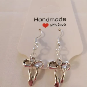 Silvriga örhängen med rosett - Handgjorda silvriga örhängen med hängande rosettformad detalj. Perfekta för att ge en söt touch till din stil. Lätta att bära och passar till många olika outfits.