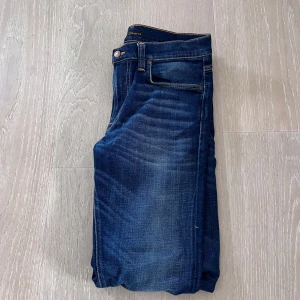 Nudie Jeans - Säljer ett par mörkblå jeans från Nudie med klassisk femficksdesign och kontrastsömmar. Jeansen har raka ben och normal passform, med läderpatch bak i midjan och diskret logga på bakfickan.