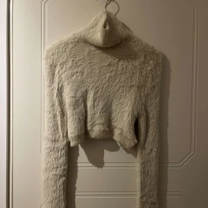 Beige fluffig croppad polotröja - Supermjuk och fluffig croppad polotröja i beige. Tröjan har långa ärmar och hög polokrage, perfekt för att sticka ut med en mysig look. Passar dig som gillar trendiga och unika plagg.