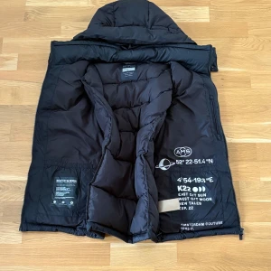  Scotch & Soda dunväst - Säljer en svart dunväst från Scotch & Soda med avtagbar huva. Västen har dragkedja och tryckknappar framtill samt tryckt text på insidan. Perfekt för lager-på-lager och har en modern, normal passform. Kolla defekter på luvan på bild 4&5. Man märker knappt defekterna.