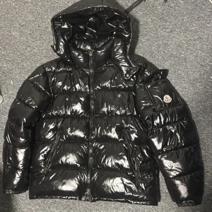 Svart glansig dunjacka från Moncler - Säljer en svart glansig dunjacka från Moncler med huva och dragkedja. Jackan har ett klassiskt Moncler-märke på ärmen och ser riktigt varm ut. Perfekt för kalla dagar när du vill hålla stilen.