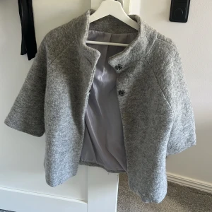 Grå ulljacka med knappar - Säljer en stilren grå jacka i ullblandning med hög krage och trekvartsärm. Jackan har två stora knappar framtill och en rak, något boxig passform. Perfekt för lager-på-lager och enkel att matcha.
