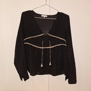 Svart blus med små prickar och knytband - Svart blus med vita små prickar och v-ringning. Blusen har långa ärmar och dekorativa knytband framtill samt volangdetalj över bröstet. Perfekt för dig som gillar en söt och avslappnad stil.