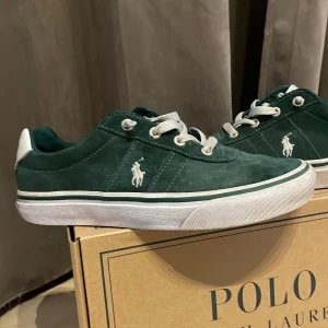 Gröna sneakers från Polo Ralph Lauren - Mörkgröna sneakers från Polo Ralph Lauren med vit logga på sidan och vit sula. Skorna har snörning och är gjorda i mocka. Klassisk låg modell med rund tå och extra skosnören medföljer. Inköpta i December 2024.