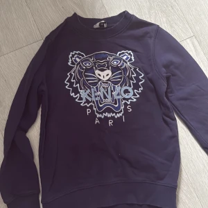 Mörkblå sweatshirt från Kenzo Kids - Säljer en mörkblå sweatshirt från Kenzo Kids med det ikoniska broderade tigerhuvudet och Kenzo Paris-loggan på bröstet. Tröjan har långa ärmar och ribbade muddar vid ärmslut och nederkant. Perfekt för dig som gillar streetwear och snygga detaljer.