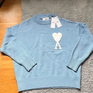 Ljusblå stickad tröja från Ami - Säljer en ljusblå stickad tröja från Ami Alexandre Mattiussi. Tröjan har en vit hjärtformad detalj med bokstaven A på bröstet, ribbade muddar och rund halsringning. Perfekt för dig som gillar stilrena och trendiga plagg. Hör av dig vid minsta fundering!