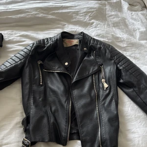 Svart skinnjacka med dragkedjor - Säljer en svart skinnjacka med coola detaljer som dragkedjor och axelpartier med sömmar. Jackan har klassisk bikerstil med snedställd dragkedja och bälte nedtill. Perfekt för dig som vill ha en edgy look.