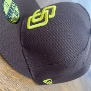 Brun keps från New Era 59FIFTY - Säljer en brun New Era 59FIFTY keps med gul broderad logga framtill och mindre broderade detaljer på sidan och bak. Klassisk flat peak och strukturerad passform. Perfekt för dig som gillar sportig stil.