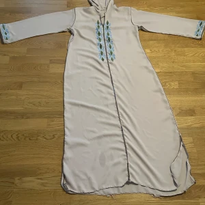 Kaftan  - Säljer en ljusbeig kaftan med huva och fina blå-gröna broderier framtill och på ärmsluten. Kaftan har långa armar och en lös passform